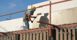 An industrial painting contractor in a hard hat and safety harness applying protective coating to the exterior wall of a commercial building using a paint roller at elevation
