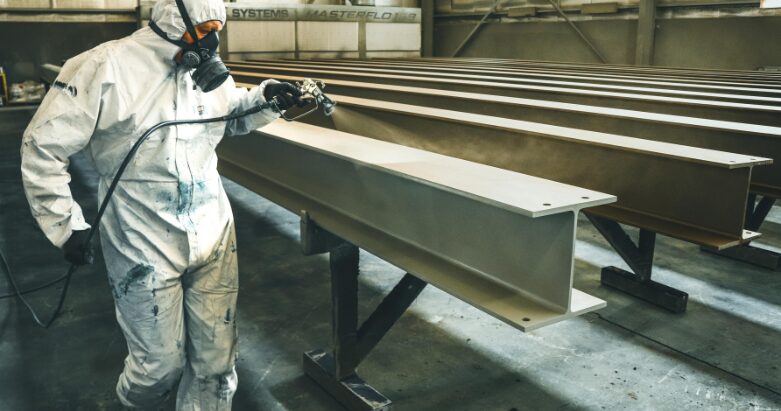 An industrial painter in a protective suit and respirator spray painting steel I-beams in a facility.