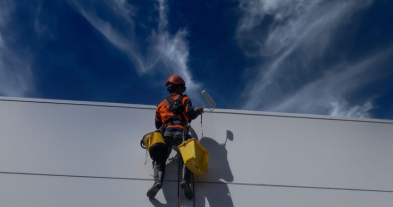 An exterior commercial painting contractor wearing a safety harness and helmet while painting a high wall under a bright blue sky.