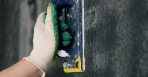 A close-up of a worker's gloved hand using a hand sander block for thorough surface preparation on a dark wall.