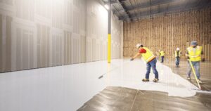 Industrial flooring contractors applying a thick light gray epoxy base coat to a large concrete warehouse floor during a professional installation.