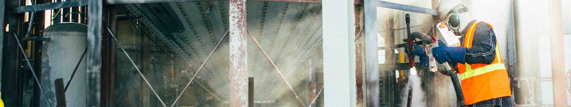 Worker in safety vest sandblasting structural steel beams at an industrial facility to remove rust and old coatings before repainting